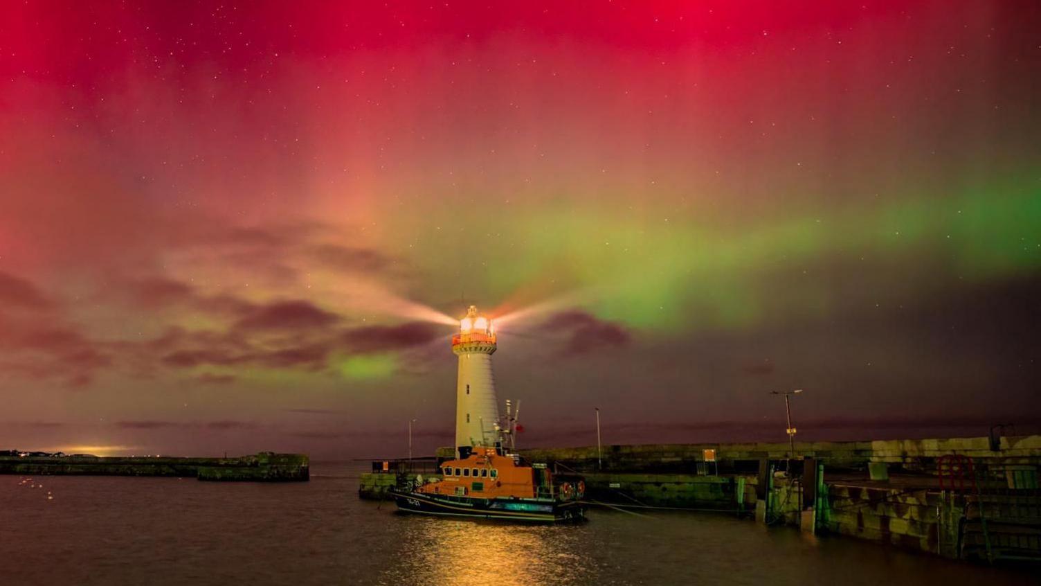 The Northern Lights lighting up the sky in pink and green colours. A white lighthouse and a harbour is at the bottom of the image. 