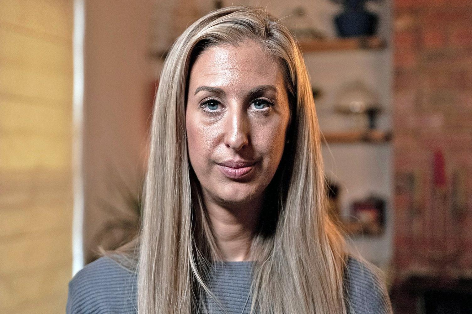 Portrait photo of Fiona Winser-Ramm. She has long, straight blonde hair with a side parting. She has blue eyes and is wearing a grey ribbed jumper. She is pictured in a living room, with shelves and a blind visible behind her. 