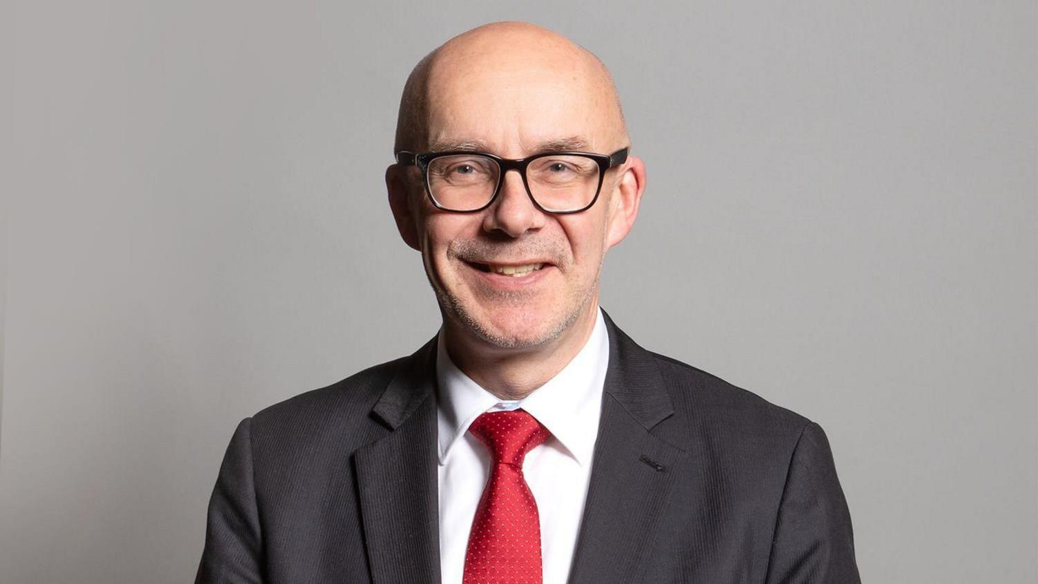 A man stands on a grey background. He is wearing a grey suit jacket, white shirt and red tie. He is bald and wearing glasses.