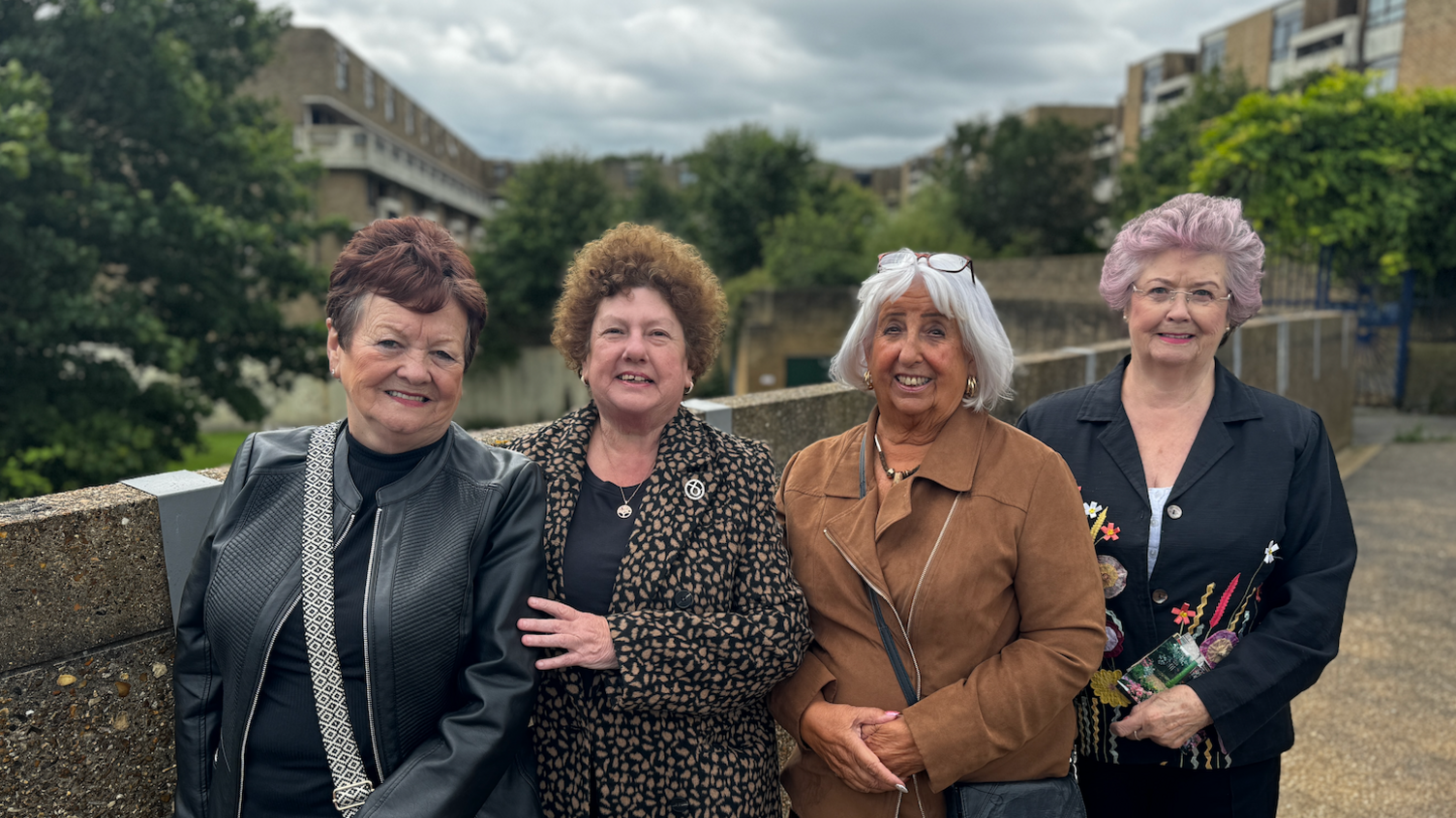 Four friends stand on a footbridge in front of flats.