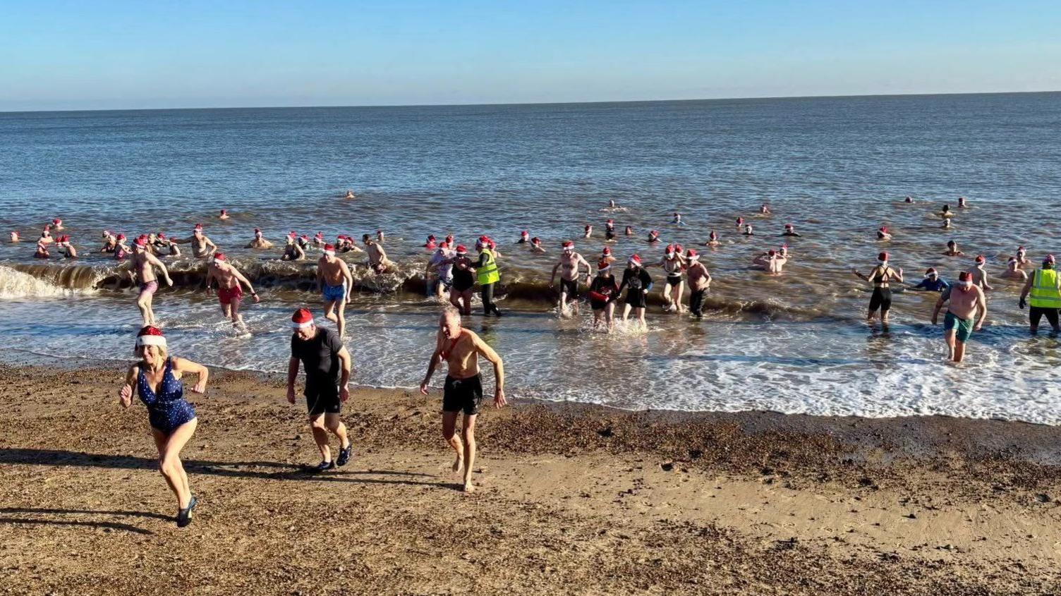 Dozens of people in the sea or returning up the beach at Aldeburgh. Many have red Santa hats on. The sea is calm and the sky is blue and clear.