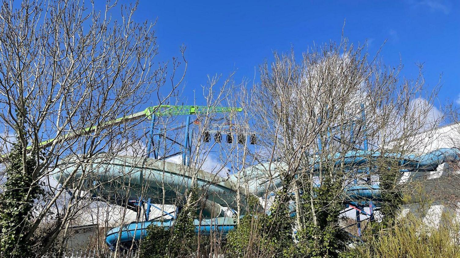 A large building with water slides coming from it behind some trees. A long green crane arm with large stage lights overhangs it all.