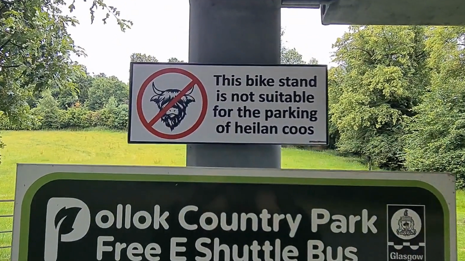 A photo from Pollok Park where Highland Cows are a popular attraction. The sign has the head of a highland cow in a circle and a line through it to show they are prohibited. The text reads: This bike stand is not suitable for parking heilan cows.