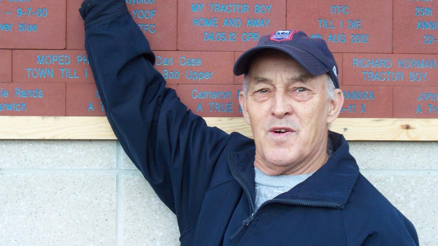 Valentine Powell, wearing an Ipswich Town baseball cap and pointing at commemorative bricks