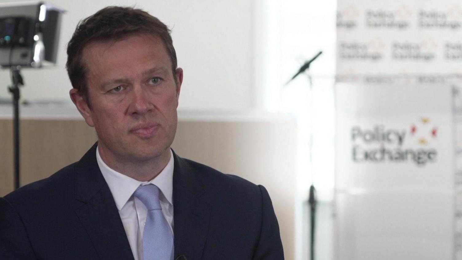 David wears a dark suit and a blue tie. He is sitting in a conference room, with the Policy Exchange logo behind him, 