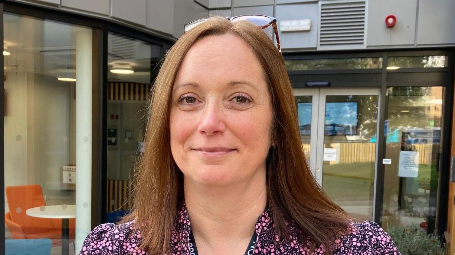 A woman with long brown hair wearing a pink and black patterned dress standing outside a building