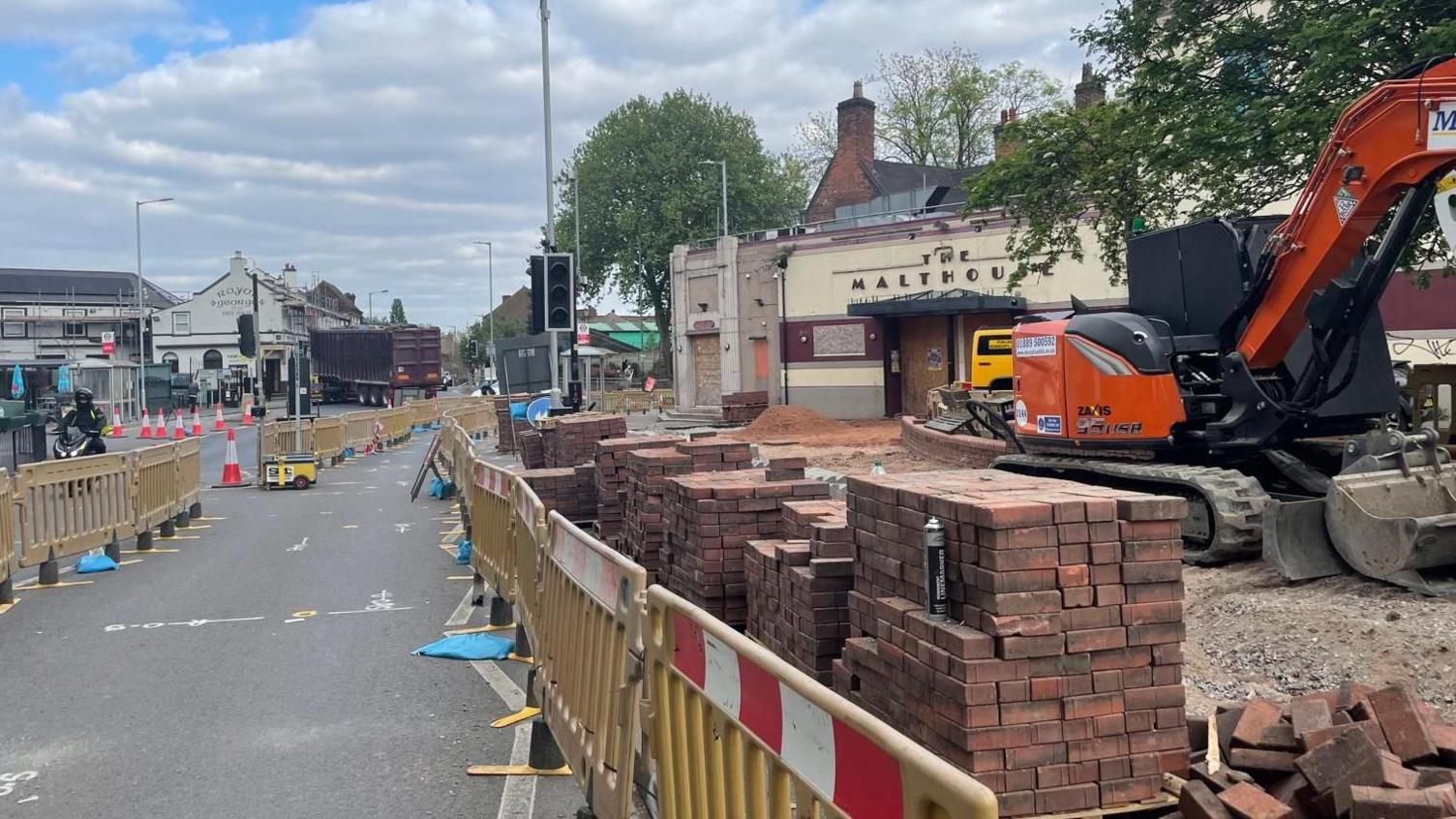 A road where there are several piles of bricks stacked up behind yellow safety barriers which have red and white markings at the top. There is an orange digger and in the distance there are traffic lights which are switched off and a series of buildings.