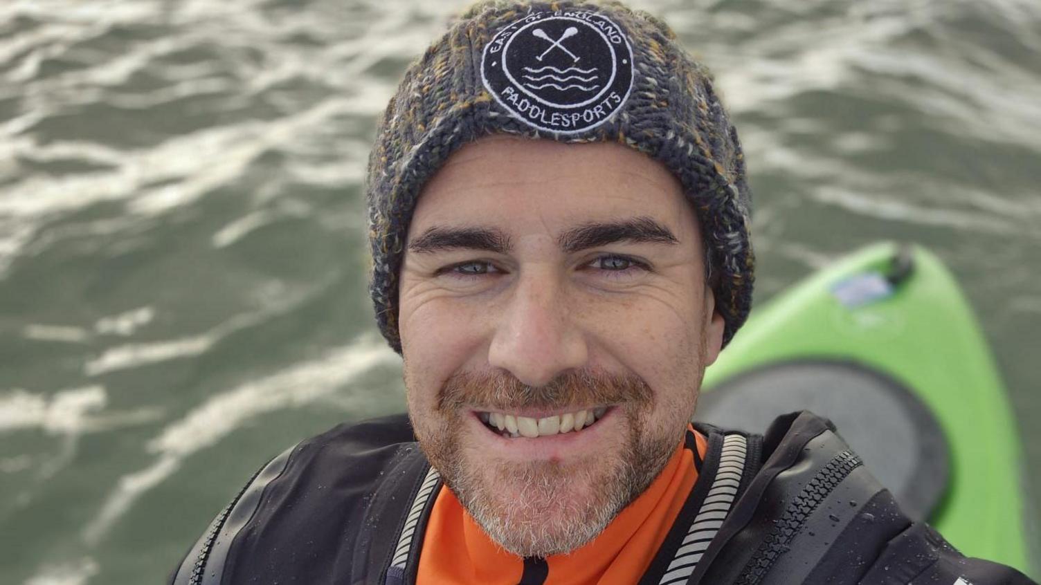 Oli Jordan has a beard, he is in a kayak and taking a selfie while smiling at the camera. He is wearing a beanie hat and a coat.