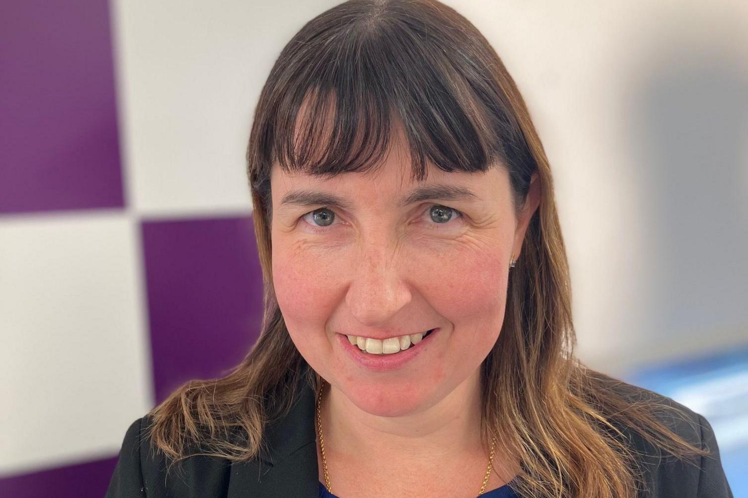 Prof Stacey Pope standing in front of the Durham University logo. She has shoulder length dark brown hair and a fringe and is wearing a blue dress, plan gold chain, and a black jacket