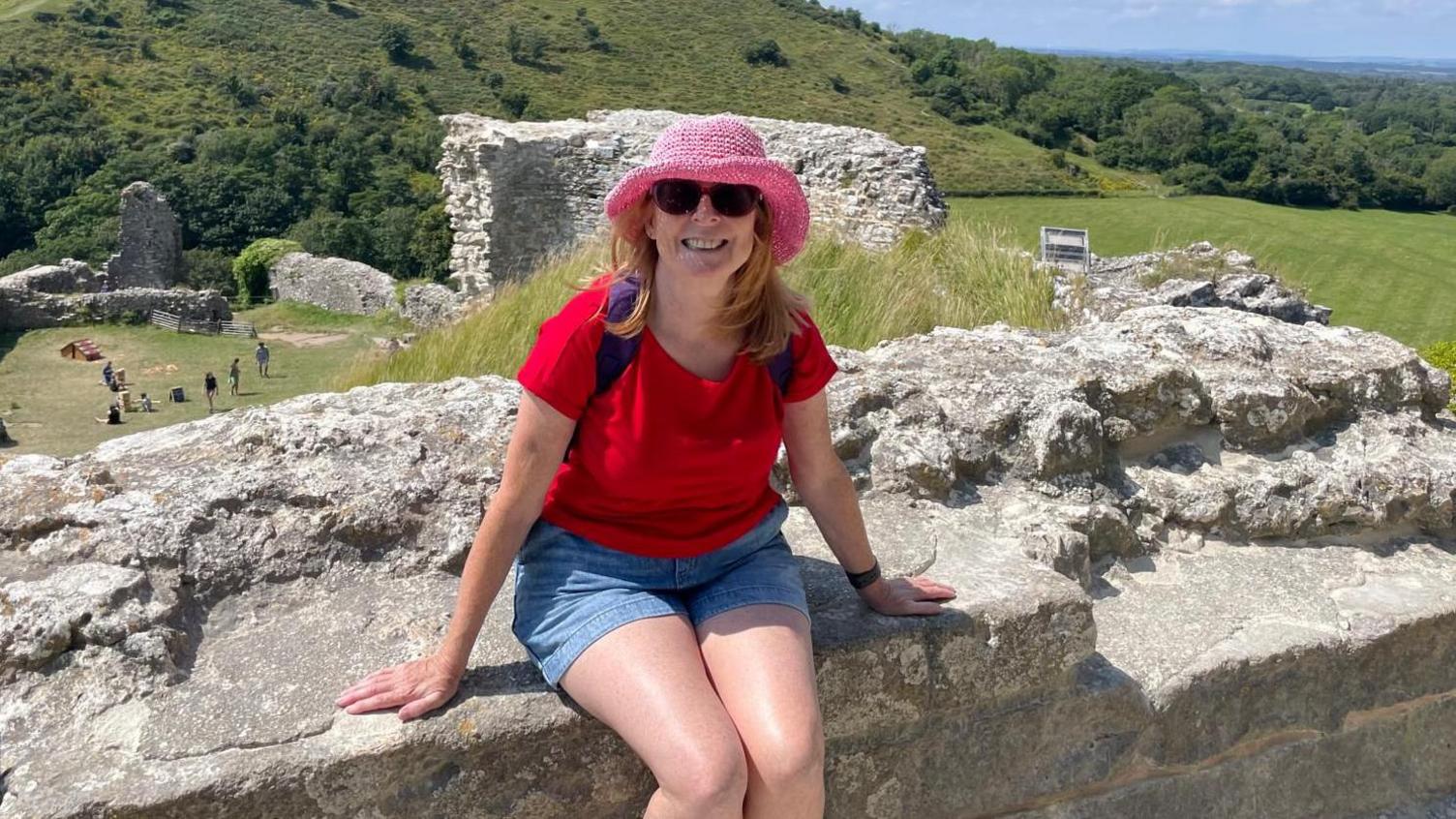 A smiling Kelly Burton wearing sunglasses, a pink hat, red T-shirt and blue shorts, sitting on a wall with grassy hills behind her