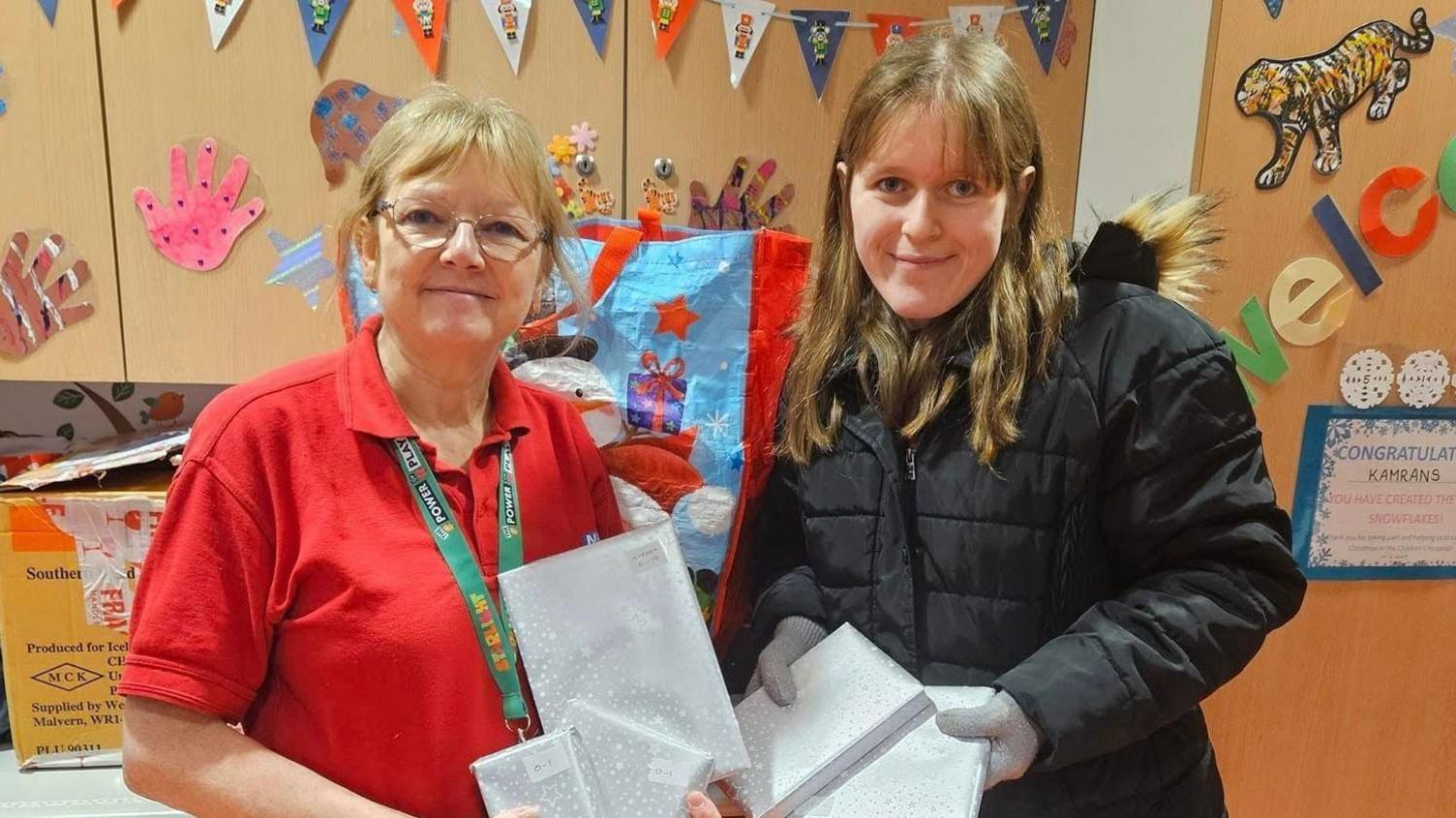 Mikayla Beames poses with an unnamed hospital ward staff member. They are both holding what appear to be Christmas presents and smiling in front of the ward's cupboards which are decorated with bunting and children's drawings.