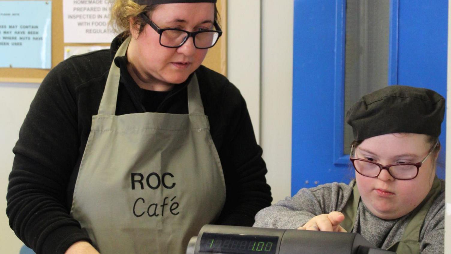 A young woman in an apron and chef's hat uses a cash till while another woman in an apron looks on