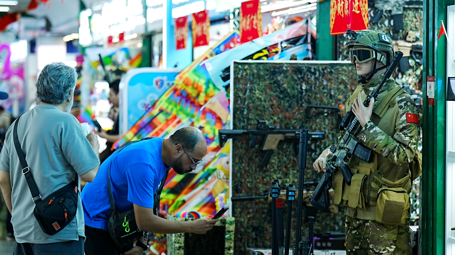 Two men look at a stall that has a life-size toy soldier standing guard, with a toy gun.