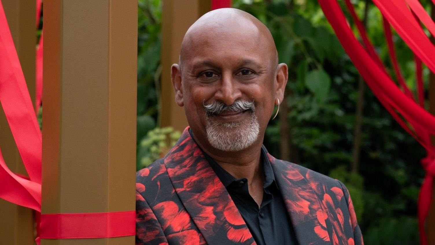 Manoj Malde surrounded by the red ribbons attached to the seating area. Th red ribbons are a symbol of HIV awareness and support. He is wearing a red and black suit, has a gold hopped earrings in his right ear and a grey moustache and goatee.