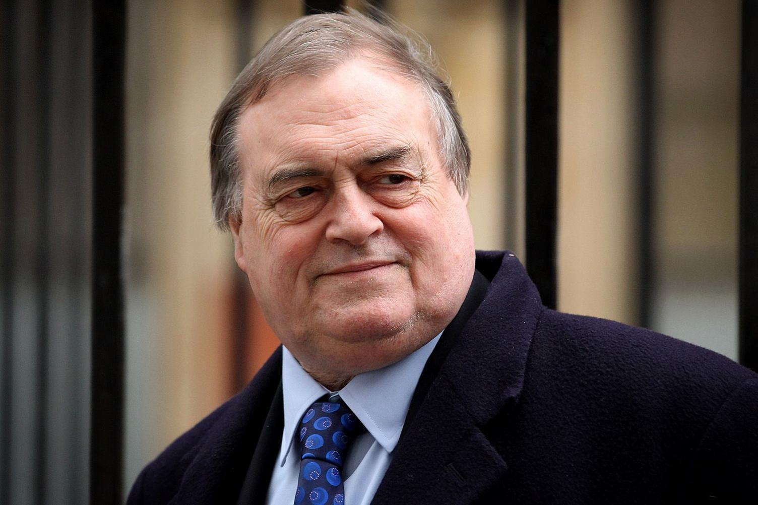 John Prescott is pictured looking slightly over his shoulder with a wry expression on his face. He has dark hair, slightly greying, and is wearing a navy blue overcoat over a suit. His shirt is light blue and his tie is dark blue.