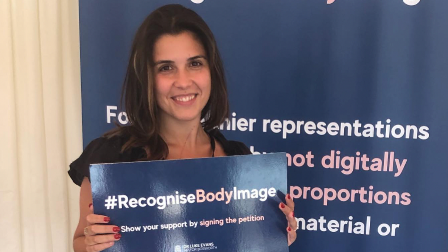 Elle Seline smiling, holding a sign that says #RecogniseBodyImage, show your support by signing the petition. She is standing in front of a banner that has wording on.