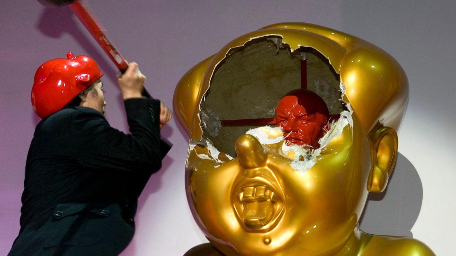 One of the Gao brothers, wearing a black suit, uses a sledgehammer to smash a gold sculpture of Mao Zedong. Inside is a red bust of Vladimir Lenin.