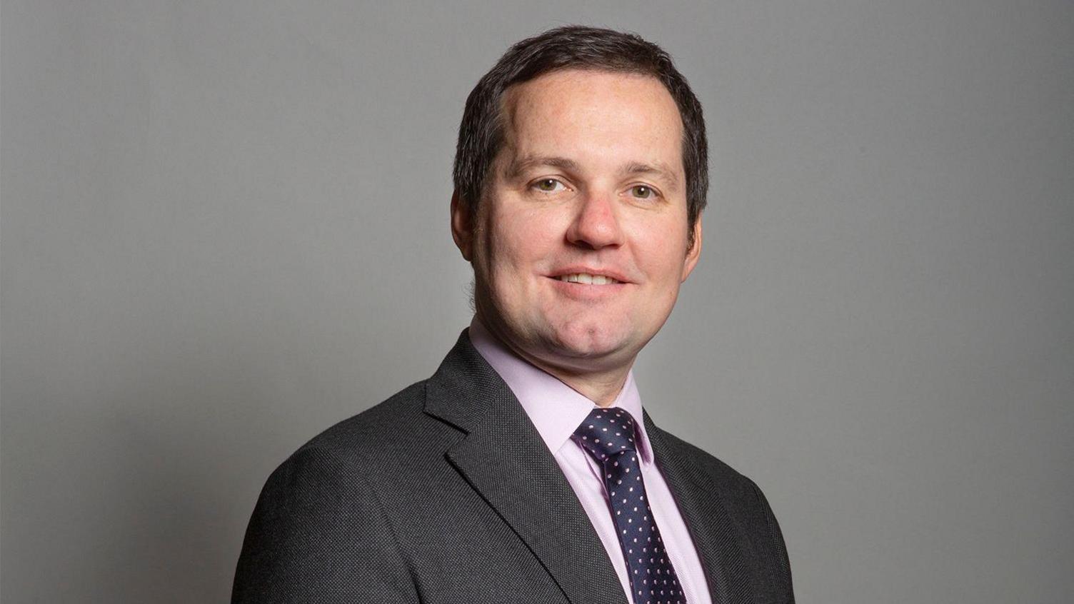 An official portrait, Chris smiles at the camera, he has short dark hair. He wears a grey suit with a pink shirt and blue tie with small pink spots