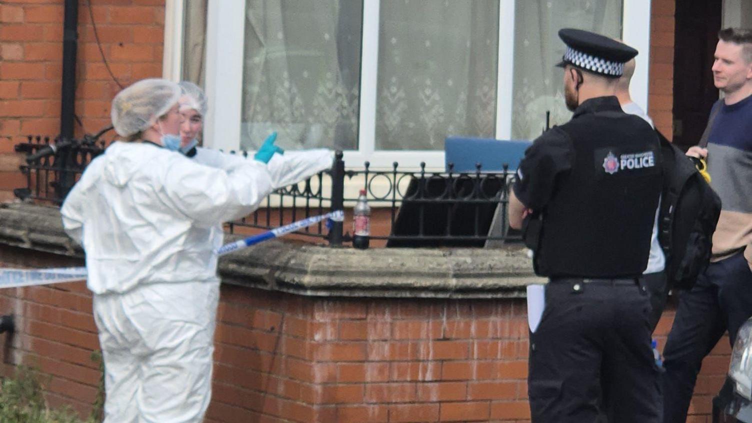 Forensic officers standing outside a house wearing white overalls and blue gloves, beside another police officer dressed in his black uniform.