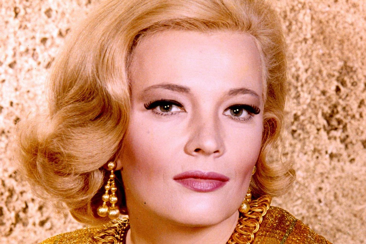 Gena Rowlands photographed against a wall in what appears to be a publicity shot, taken around 1960 when she was 30. She is wearing make-up and staring straight at the camera, she has carefully styled shoulder-length blonde hair and has gold earrings which (from what little is visible) appear to match the colour of her dress. 