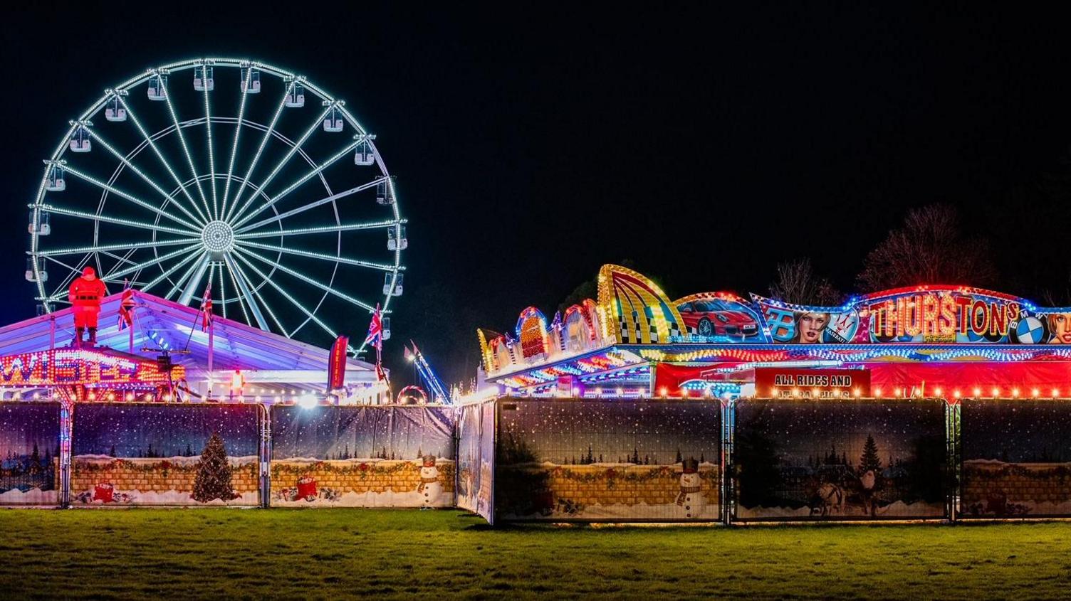 A view of a fairground from a distance at night. There are stalls in front with a ferris wheel lit up in the background.