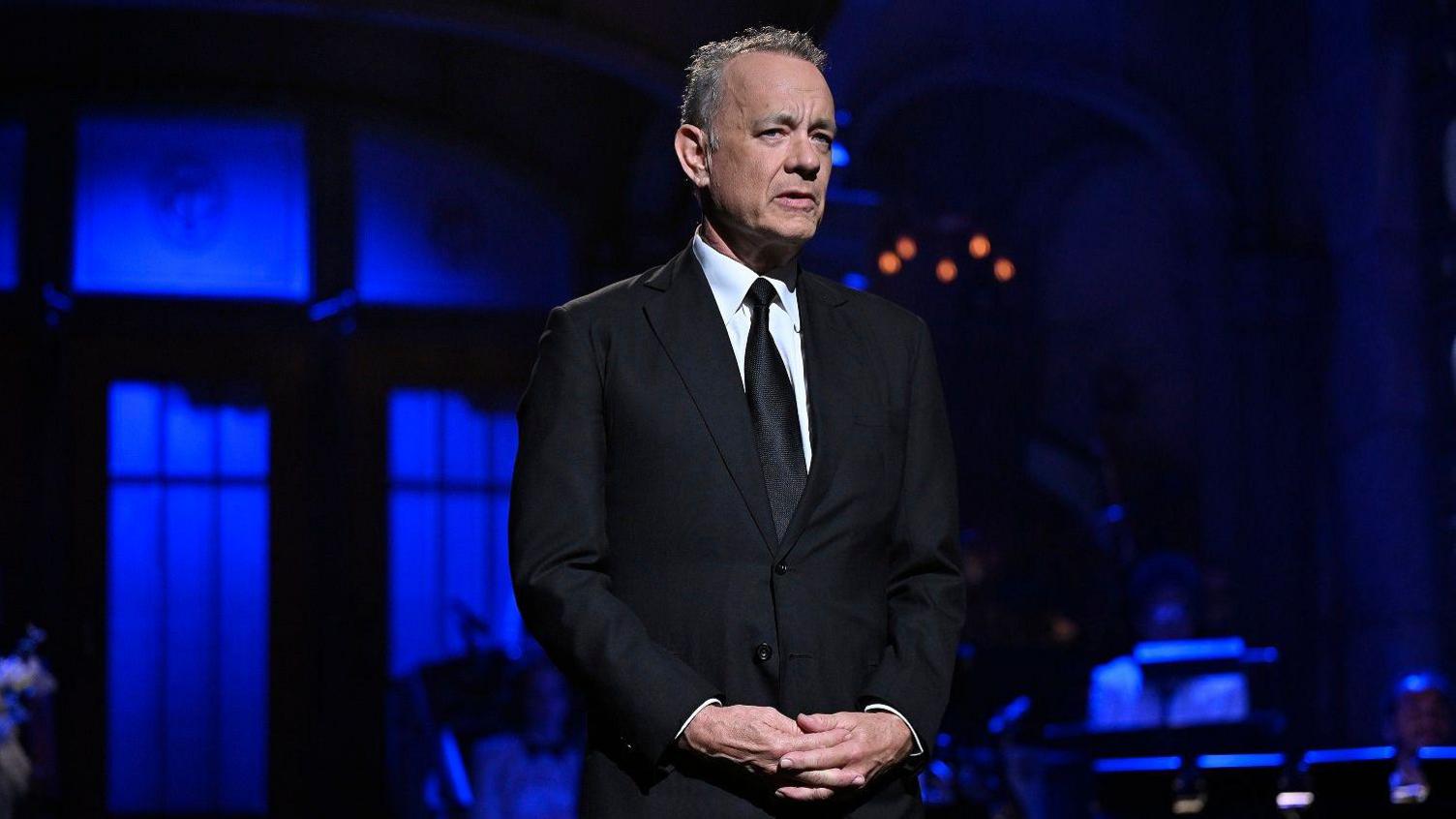 Tom Hanks interlocks his hands infront of him wearing a black suit and tie with a white shirt