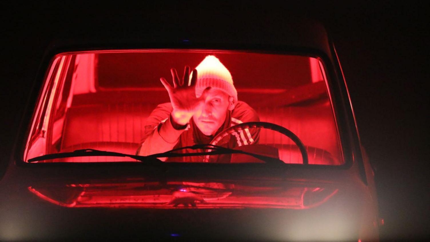 Marc Brew sat in a car on stage with red light illuminating the car