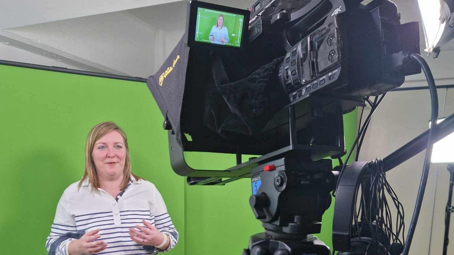 Milly Fyfe has long blond hair and is wearing a blue and white top and is standing in front of a green screen and talking to a camera