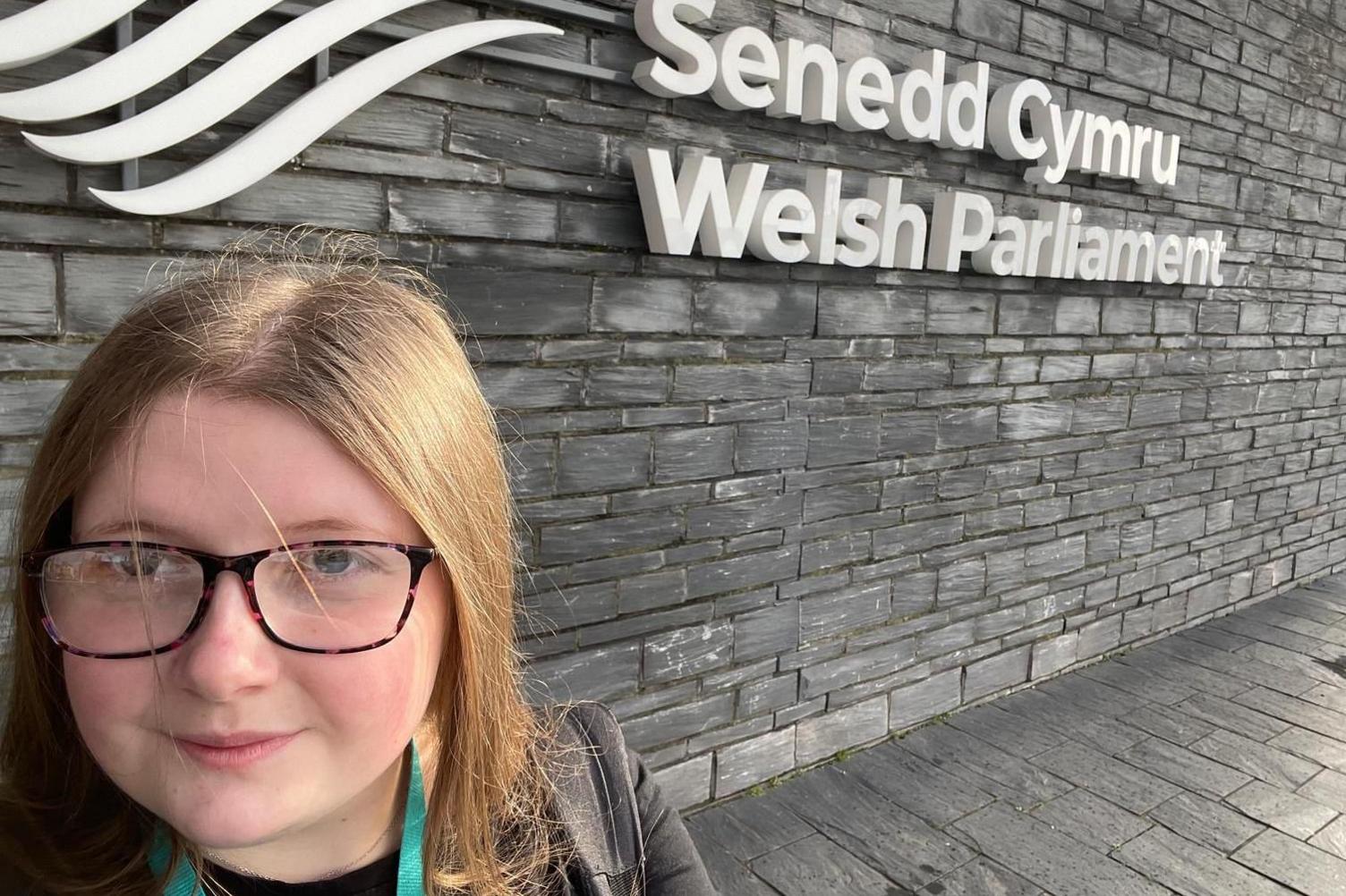 Ffion Haf Scott o flaen arwydd 'Welsh Parliament'.