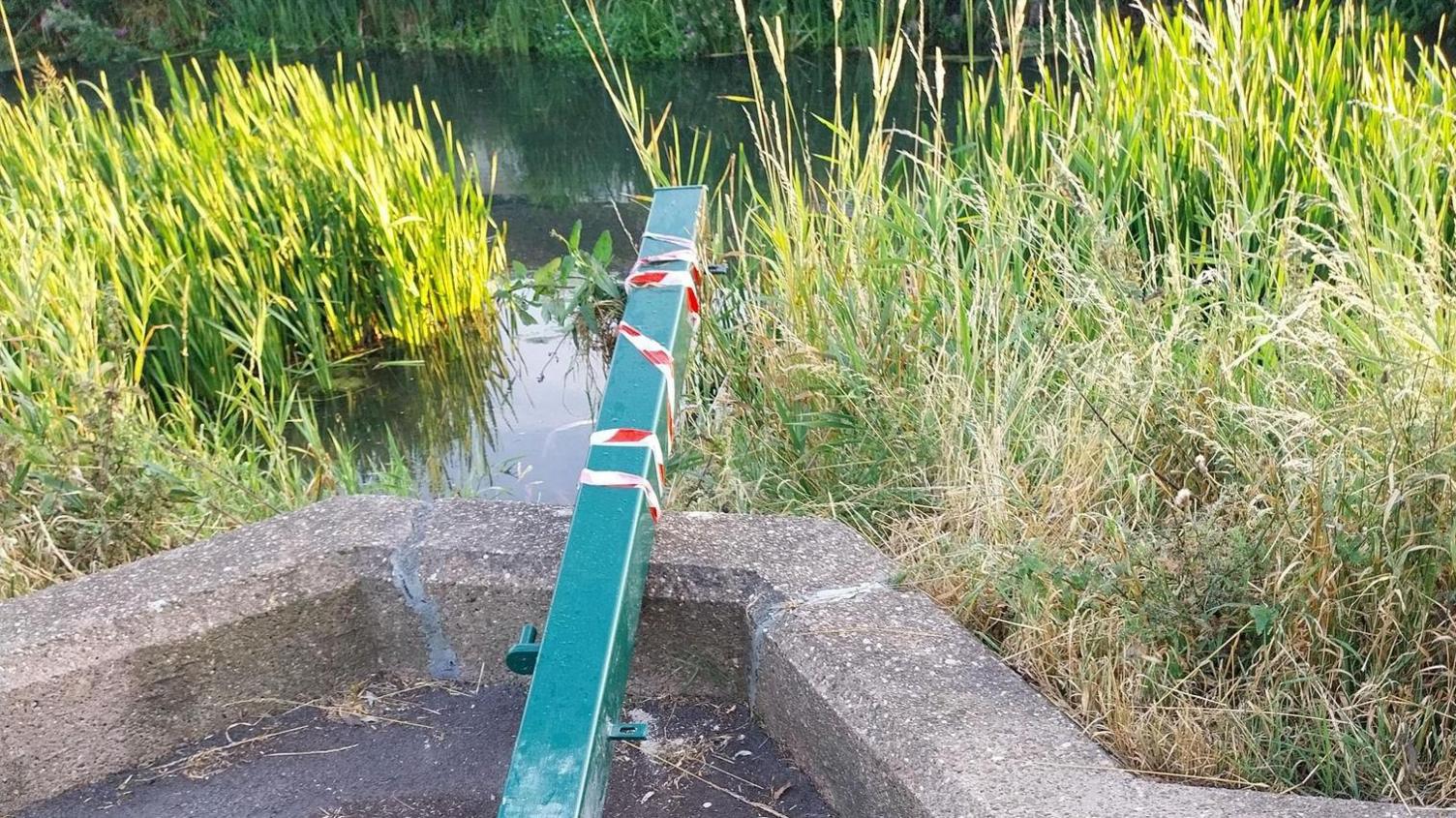 A fence post by a river. It is turqoise in colour and has red and white tape wrapped around part of it. 