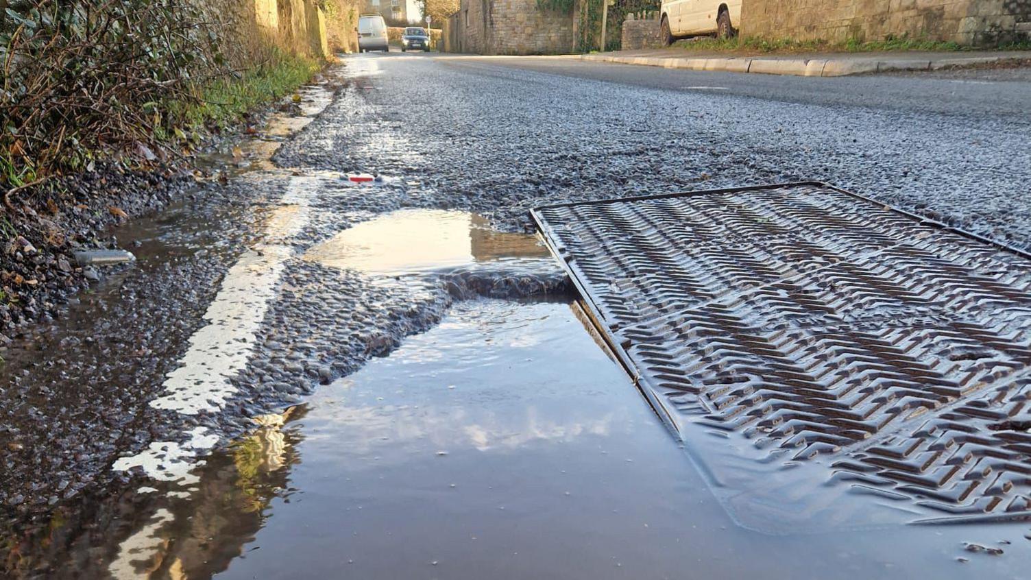 Water in massive hole in the road. It's on a narrow road with a three giant drain covers next to it.