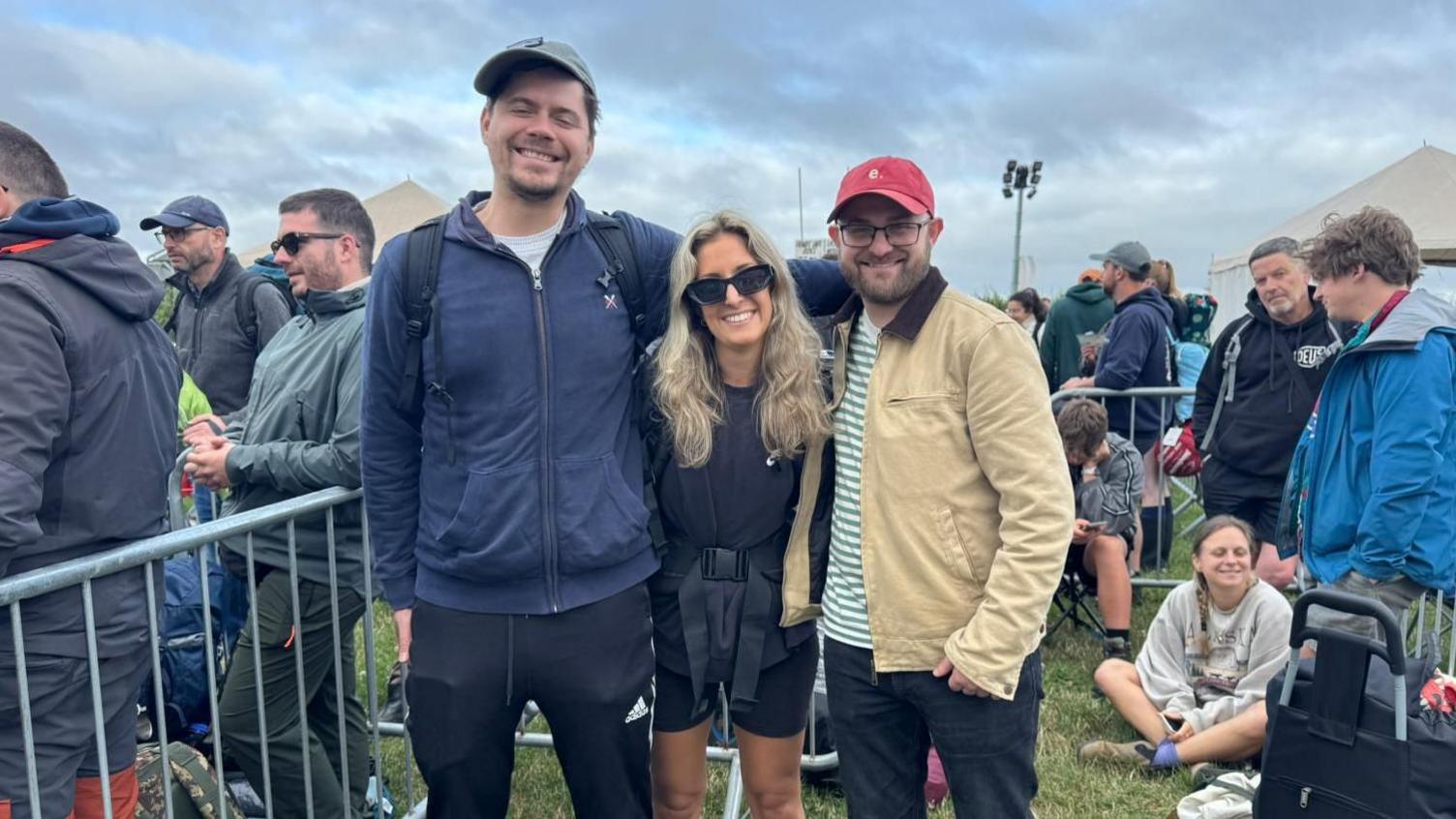 Three people standing by the line to enter Glastonbury Festival. There is a man dressed in a blue zip up jacket who is wearing a cap. He has his arms around a blonde woman who is dressed in all black and wearing sunglasses. Another man is stood next to her. He is wearing a green and white striped t-shirt, a beige jacket and black trousers with a red cap. They are all looking at the camera and smiling. There are crowds of people behind them waiting to enter the site.