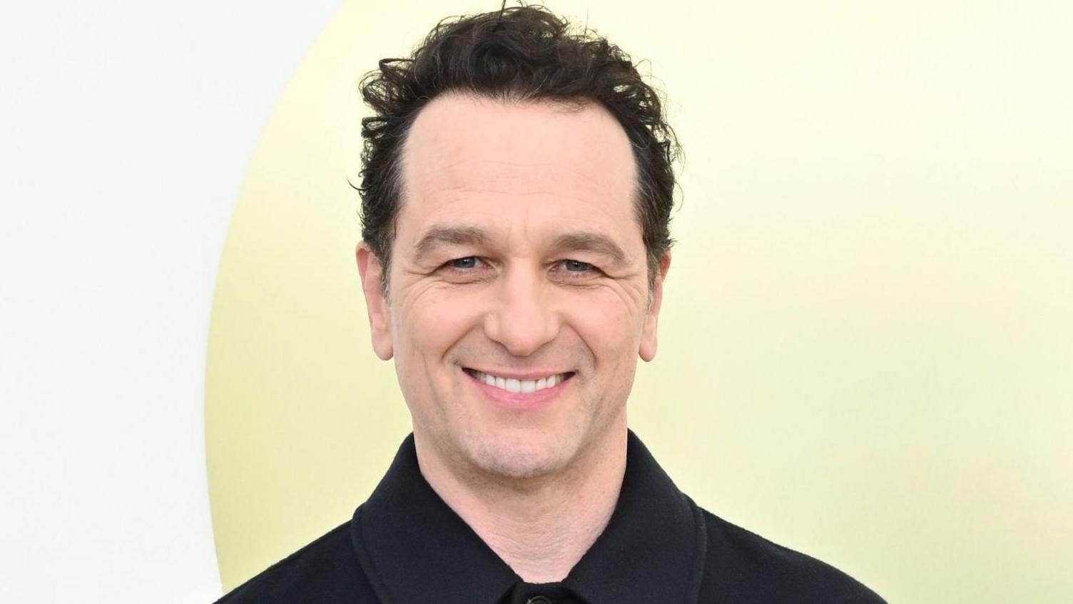 Rhys on a red carpet, wearing a black shirt. He is clean-shaven with short dark hair, and is standing against a pale yellow backdrop. 