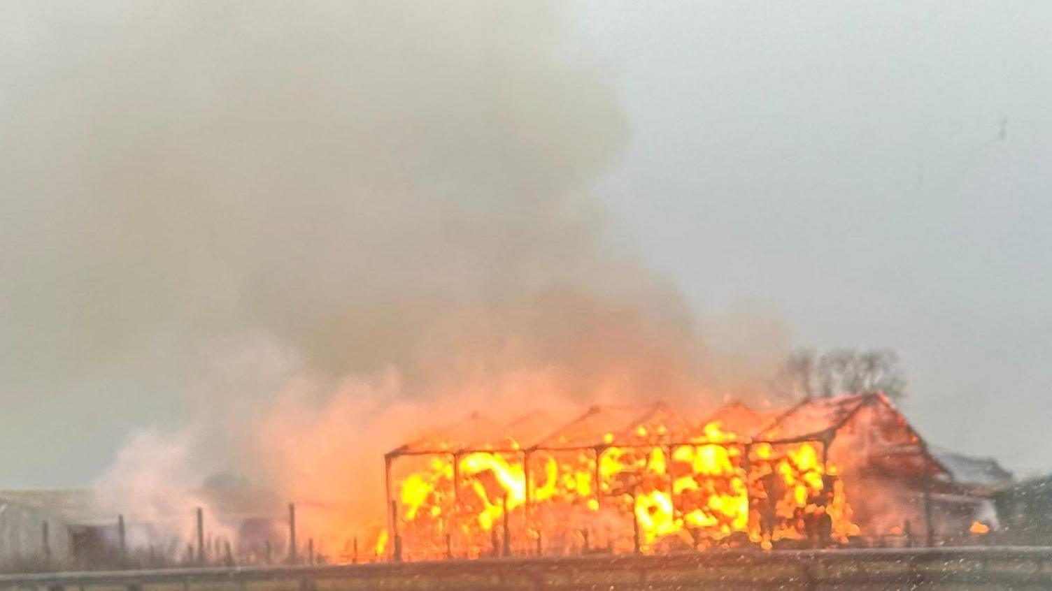 A picture taken from the A1 of the fire. The barn is engulfed in orange flames. Only the framework of the building remains standing. A large plume of smoke trails into the air.