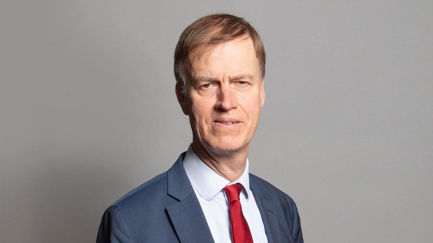 Minister for disability health and work, Sir Stephen Timms looks at the camera in front of a plain grey background. He is wearing a red tie to represent his position in the Labour party and is wearing a grey suit with a pin striped, white shirt. He has brown hair swept to one side.