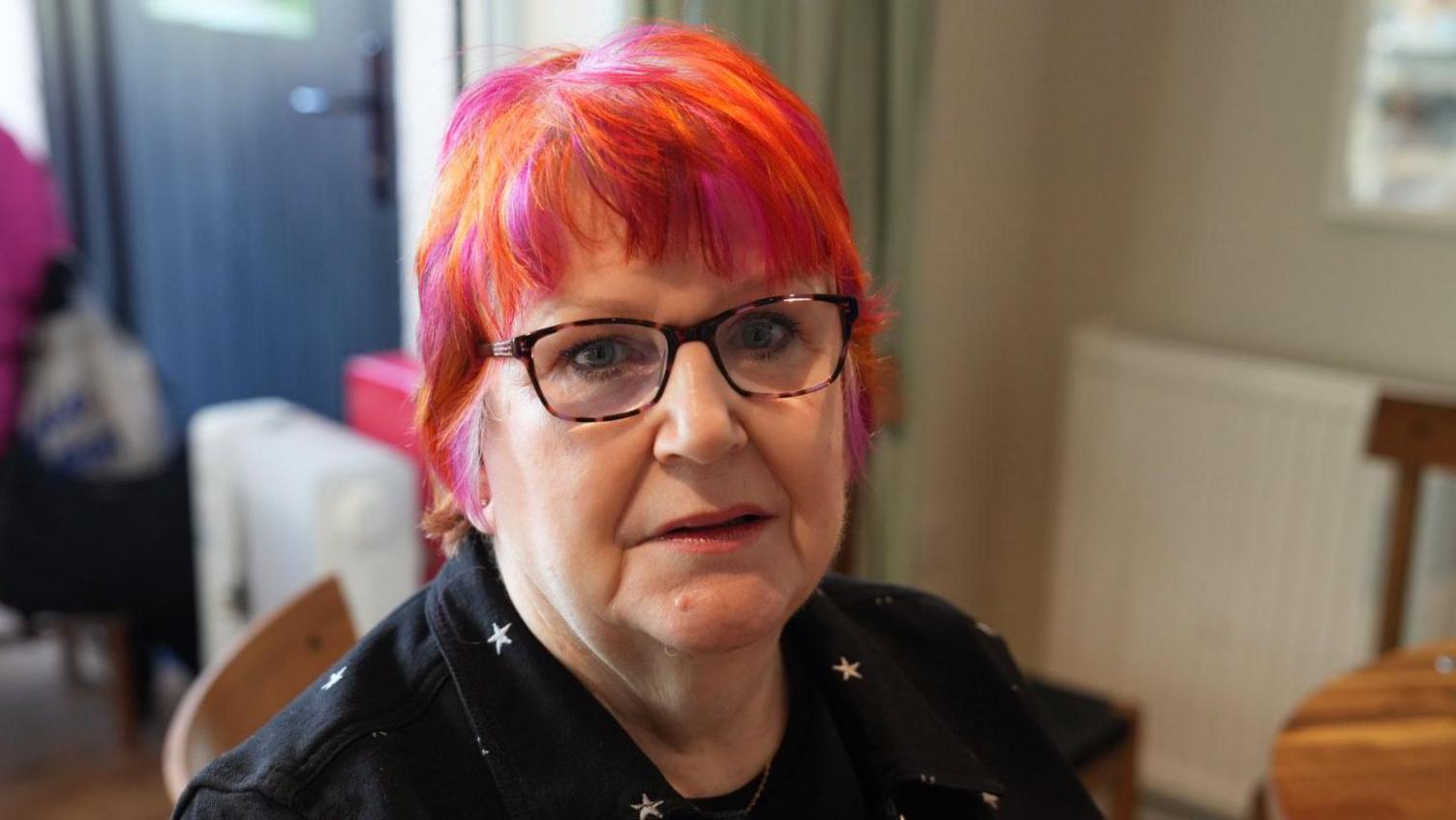 Avril Green wears a black shirt with silver stars. She has tortoiseshell glasses and cropped hair which is pink and orange.