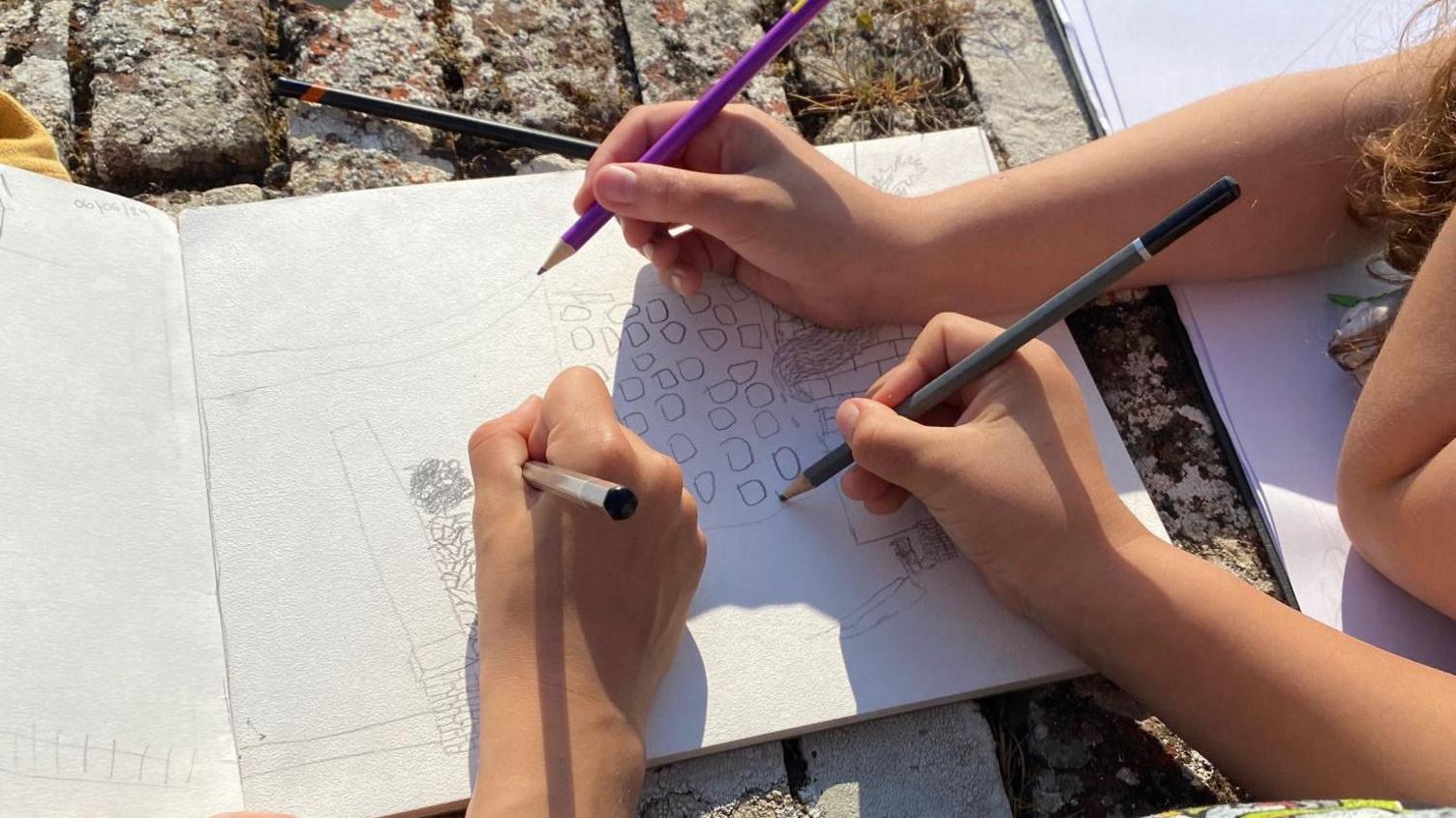 Three hands with pencils in their hands drawing a sketch in a sketchbook on the ground.