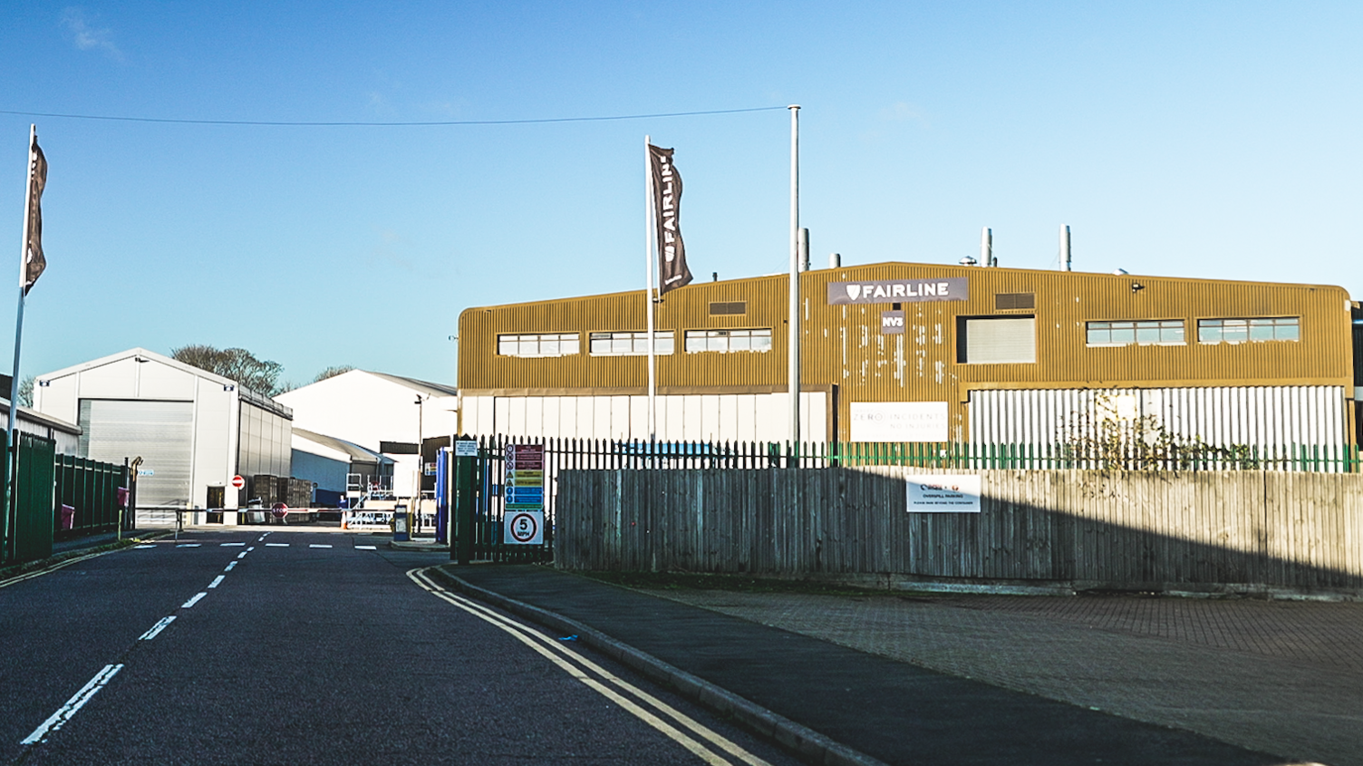 A yellow and white corrugated iron building surrounded by a green metal fence. There is a road in the foreground, with double yellow lines painted on it.