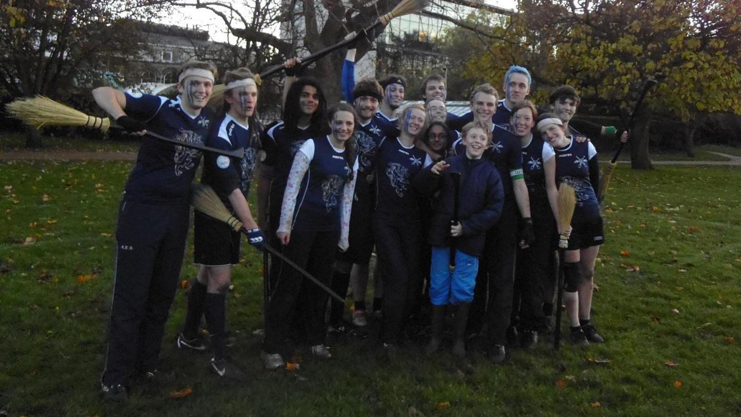 A group of people stand in front of a tree. Some hold broomsticks, others wear headbands. A young boy in blue trousers stands in the centre of the group, smiling.