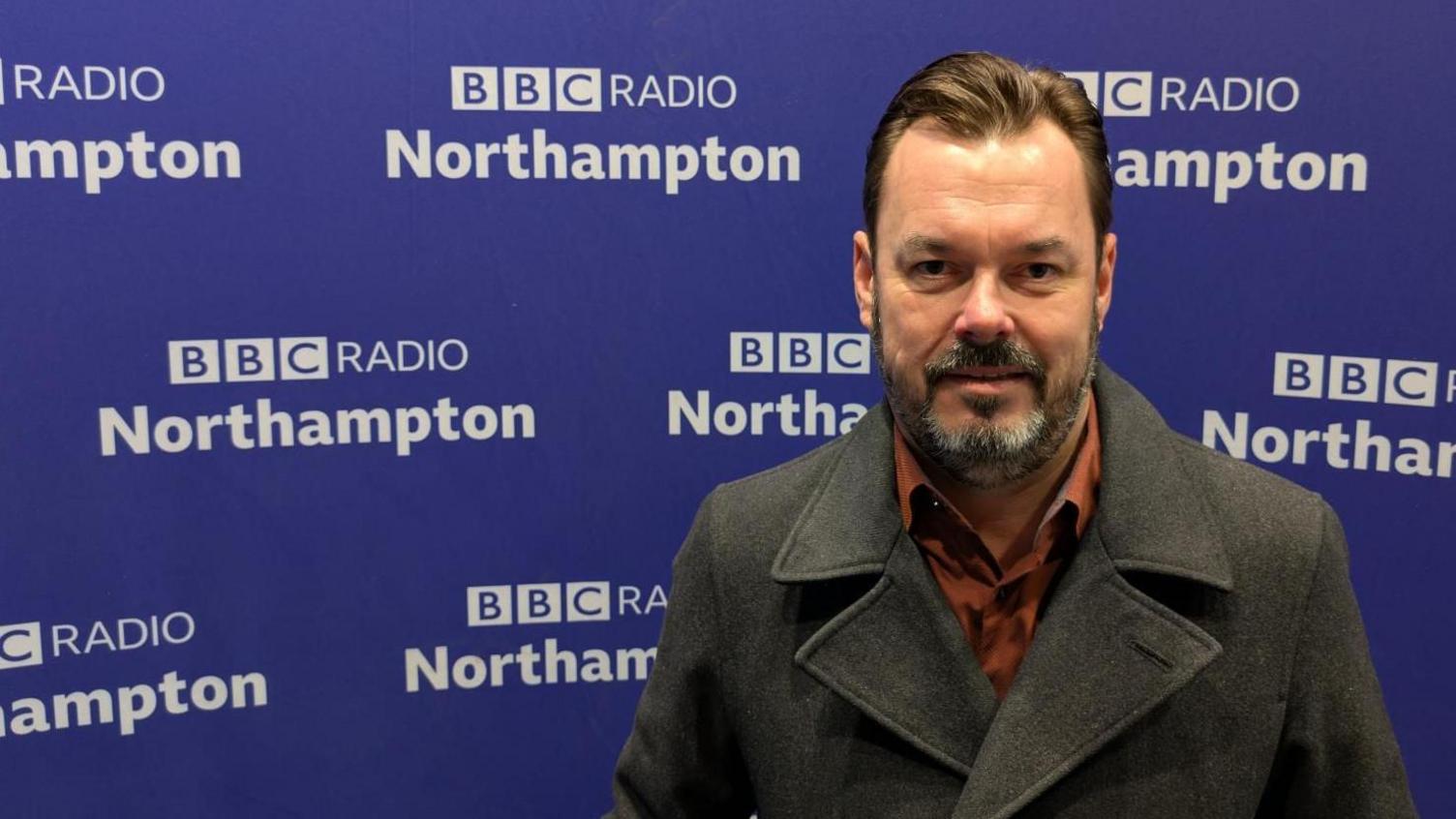 A man in a burnt orange shirt and a charcoal pea coat stands in front of some purple BBC Radio Northampton branding. 