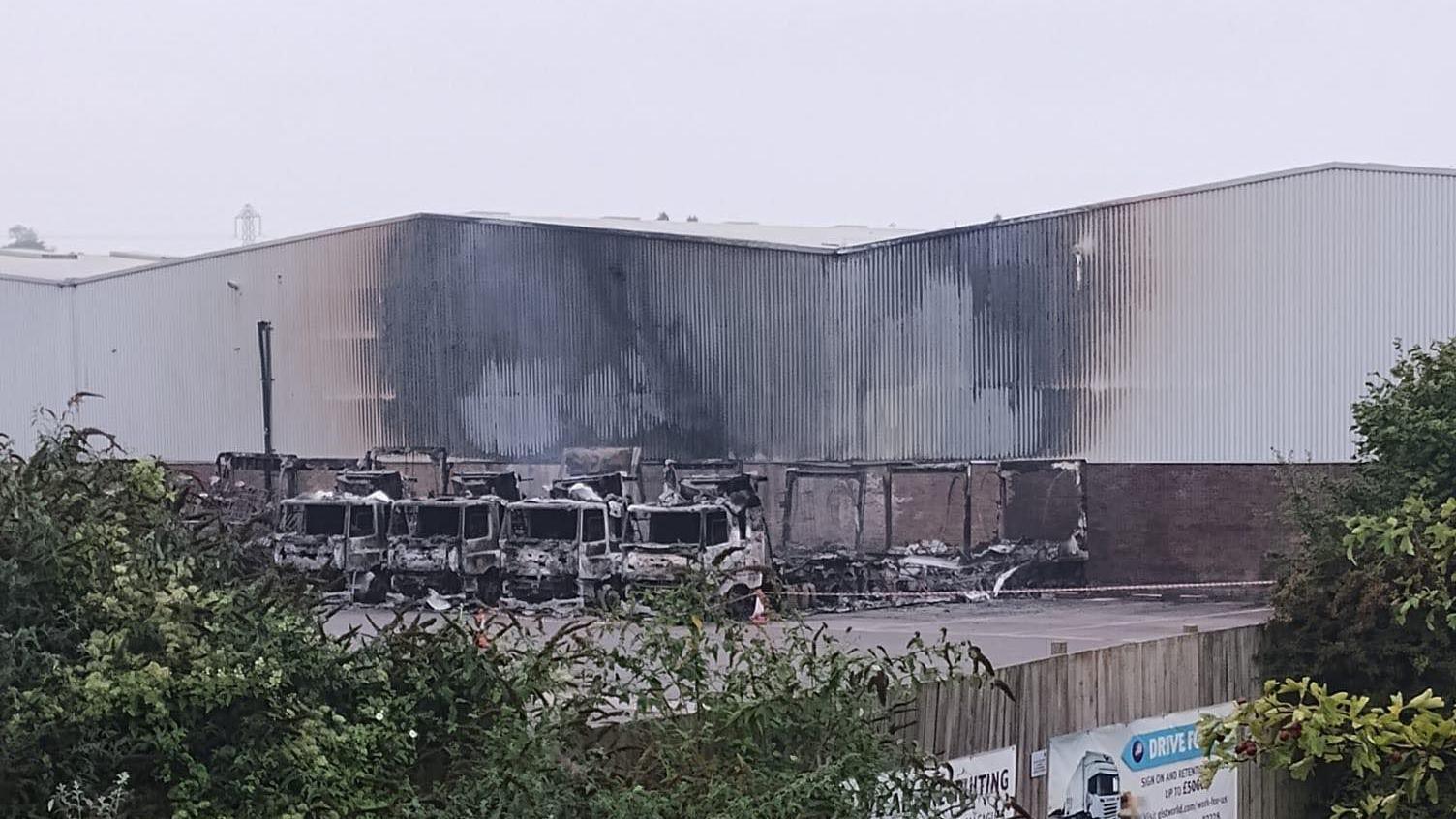 Burnt up lorries parked outside a warehouse. The side of the building is black from the fire and the lorries are badly damaged. 