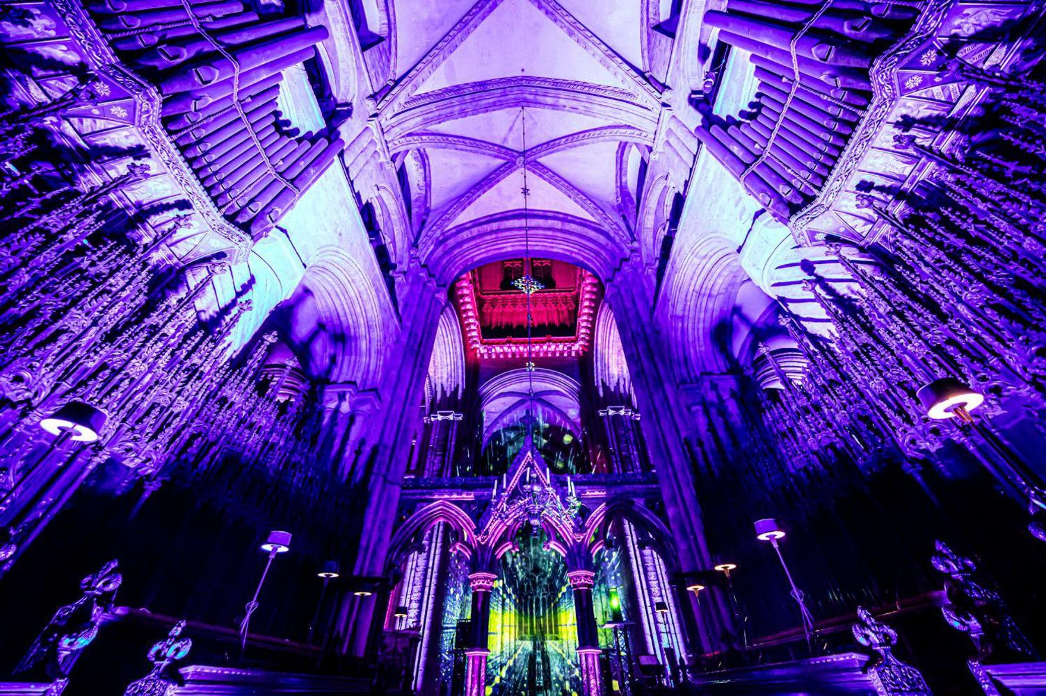 The cathedral nave is lit up in purple with green projections. Parts of the columns and the ceiling are lit up in different shades of pink and red. 