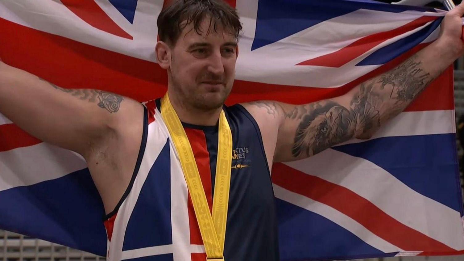 Ben on the podium with a gold medal around his neck holding a Union Jack flag.