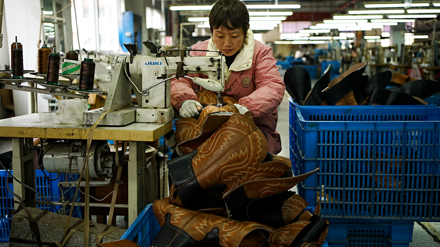 A worker at the factory sits at a wooden bench with an old sewing machine on it, sewing brown leather cowboy boots. She is wearing a pink and white jacket, and there are large blue containers next to her full of boots.