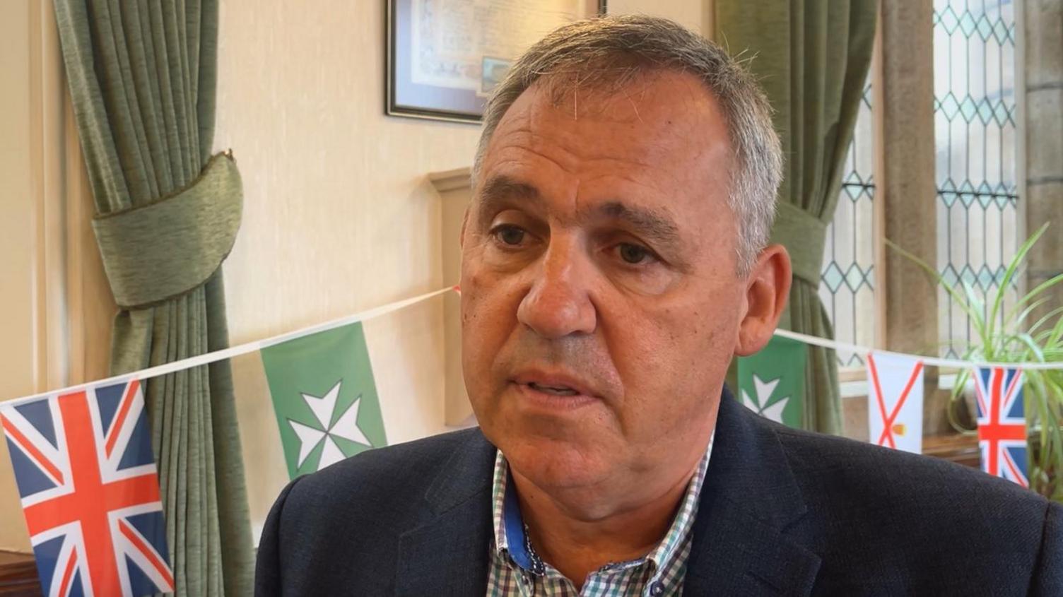 Andy Jehan pictured in St John's parish hall. There are curtains and windows behind him, along with Jersey and St John's parish flag bunting. Andy is wearing a blue blazer and check shirt.