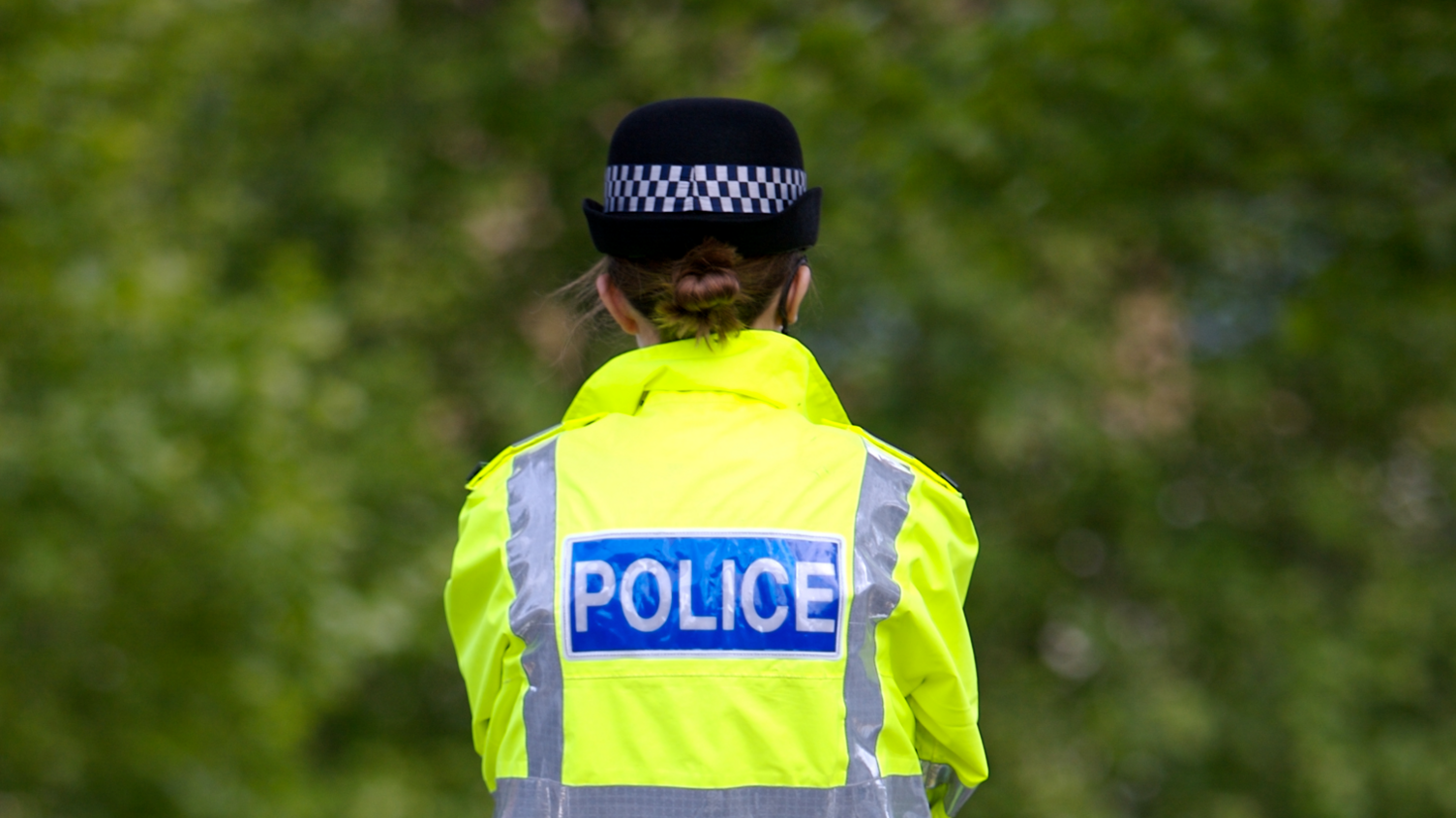 A female police officer seen from behind wearing a yellow police jacket and standing in front of some trees