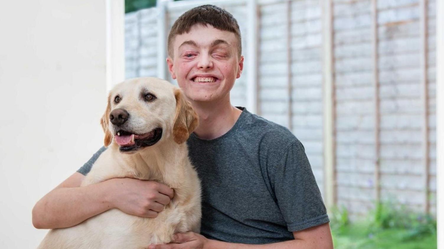 Declan sits at home, wearing a dark t-shirt, and hugging a golden Labrador, with the lawn and garden fence visible through the window behind him.
