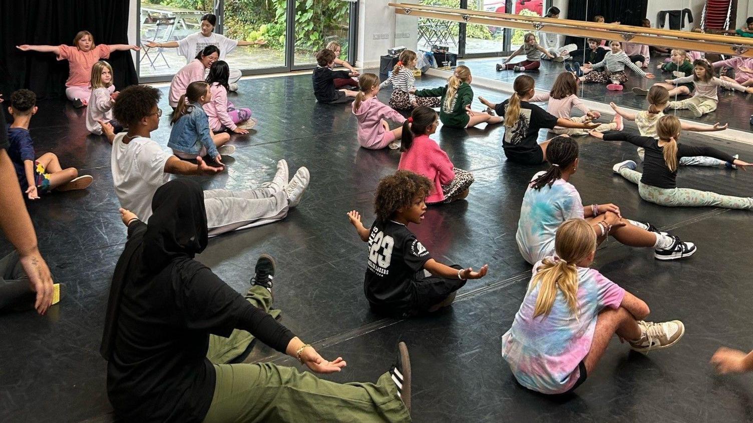 A dance studio with children sitting on the ground in front of a mirror as they are being shown dance moves. An outside area could be seen through the glass doors.