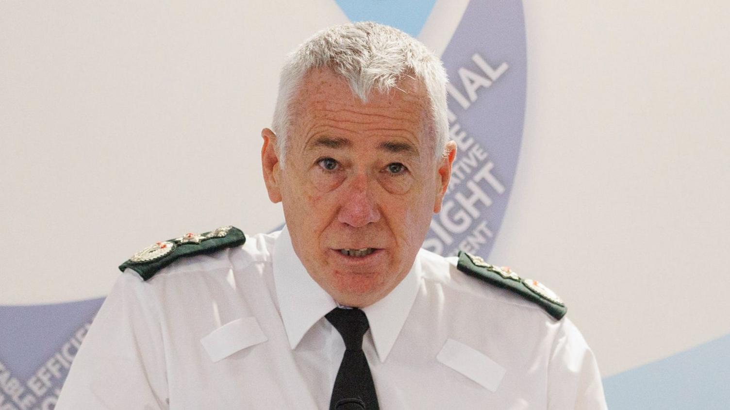 PSNI Chief Constable Jon Boutcher speaking during at a lectern. He has white hair and wears a white shirt and police shoulder lapels. 