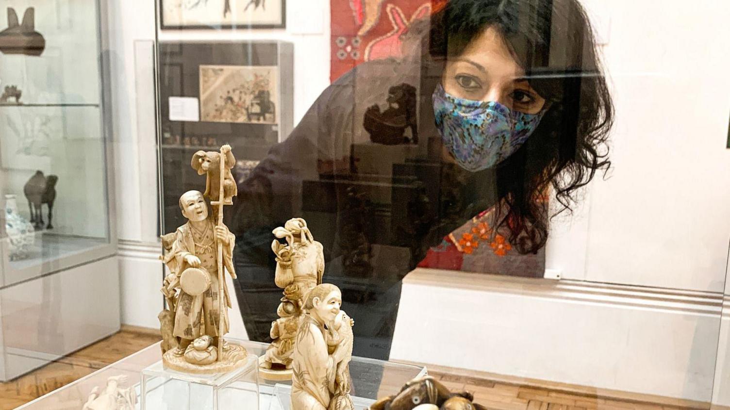 A woman with black hair and wearing a black top and a blue patterned face mask, looking at a small statue inside a glass case.